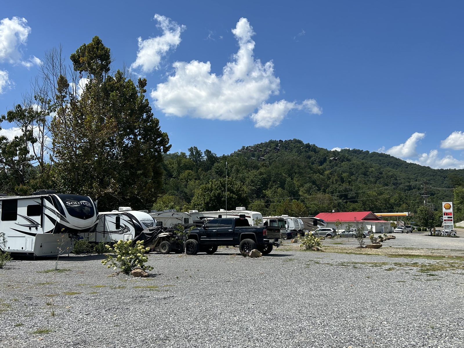 RVs parked on gravel lots with a wooded mountain behind and the Shell station next door